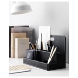 Black desk organizer with compartments for pens, markers, and ruler. Made of laminated paperboard. Surrounded by books, lamp, and papers.