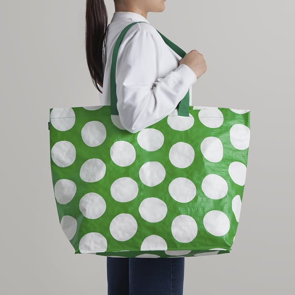A person carries a green shopping bag with white polka dots, designed to be easily cleanable and storable, with versatile carrying options.