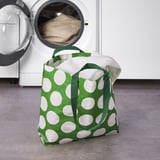 Green polka dot laundry bag, filled with white towels, stands upright near open washing machine.