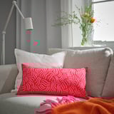 Bright red patterned pillow on grey couch with white pillows, near window with flowers.