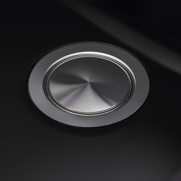 Round stainless steel sink drain cover on black sink. High-quality, reflective surface with smooth finish. Centered, polished metallic design.