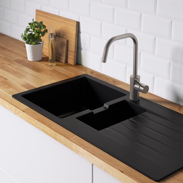 Black kitchen sink with drainboard, stainless faucet, wooden countertop, white brick backsplash, bottle, plant, cutting board.