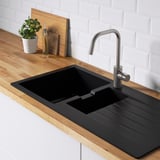 Black kitchen sink with drainboard, stainless faucet, wooden countertop, white brick backsplash, bottle, plant, cutting board.