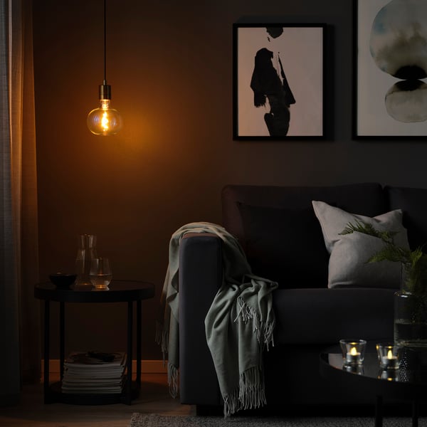 Dim room with SKAFTET lamp over table. Gray sofa with blanket, pillows, and wall art.