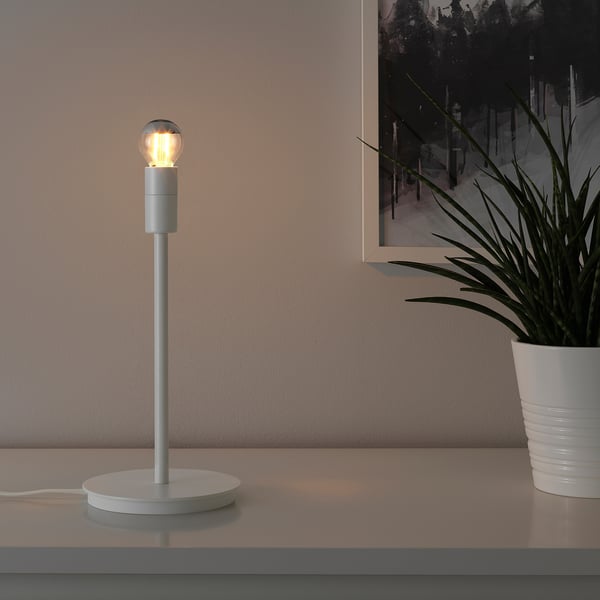 A modern lamp with a turned-on bulb on a white table. Beside a potted plant and a picture frame.