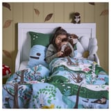 A child sleeps on a forest-themed bed, hugging two soft otter toys. The bedding and a wall decoration feature woodland motifs.