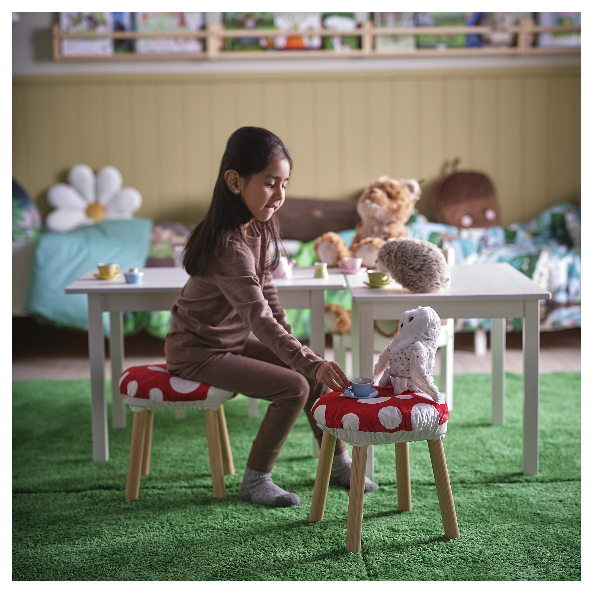 Heavy brown-haired girl sits on red polka dot stool playing tea party with stuffed animals. Bright, cheery room with green rug and white furniture.