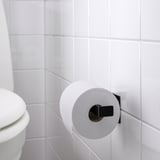 Black toilet paper holder mounted on white tiled wall, holding a roll of white toilet paper, near a white toilet.