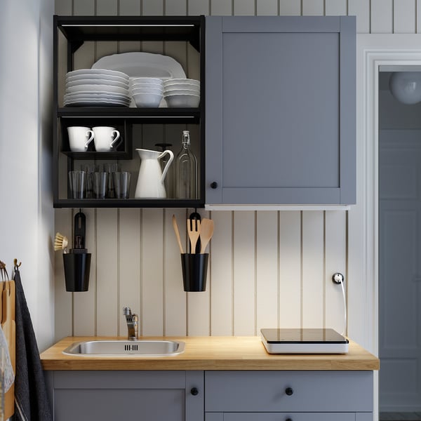Kitchen workspace with sink, grey cabinet, wooden counter, black utensil holders, and open shelf with plates, cups, glasses, and jars.