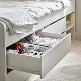 Open white bed drawer filled with neatly folded colorful clothes under a made bed.