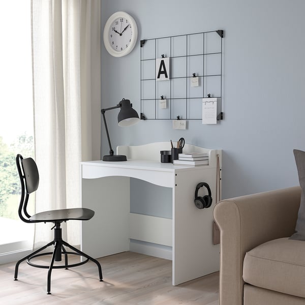 Small white kids desk, SMÅGÖRA, with black office chair. Includes lamp, headphones, books. Grey wall, clock above.