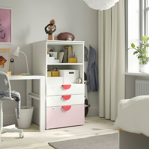 A white SMÅSTAD bookcase with pink drawers, storing various items in a room. The bookcase stands near a table with a lamp and a window with curtains.