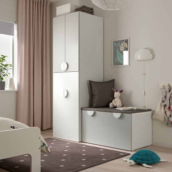 Childs room with white SMÅSTAD wardrobe, toy box, rug, and crib. Soft lighting, curtains, and wall art.