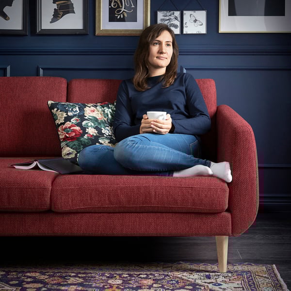 A woman sitting on a red SMEDSTORP sofa, holding a cup, with dark blue walls and floral cushions.
