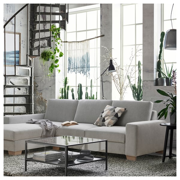 Modern living room with grey SÖRVALLEN sofa, glass coffee table, industrial staircase, large windows, and various green plants. Minimalist design.