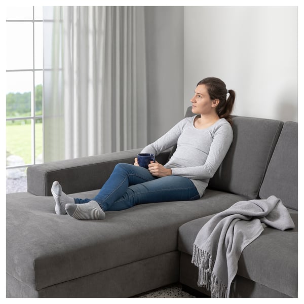 Woman relaxes on grey SÖRVALLEN sofa, holding coffee, with large window and drape behind.