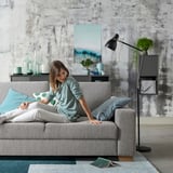 Woman relaxes on grey SÖRVALLEN sofa, minimalist design, textured fabric, decorative throw pillow, modern living room with abstract art.
