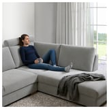 Woman relaxes on grey SÖRVALLEN sectional sofa, holding a mug, near large windows with curtains.