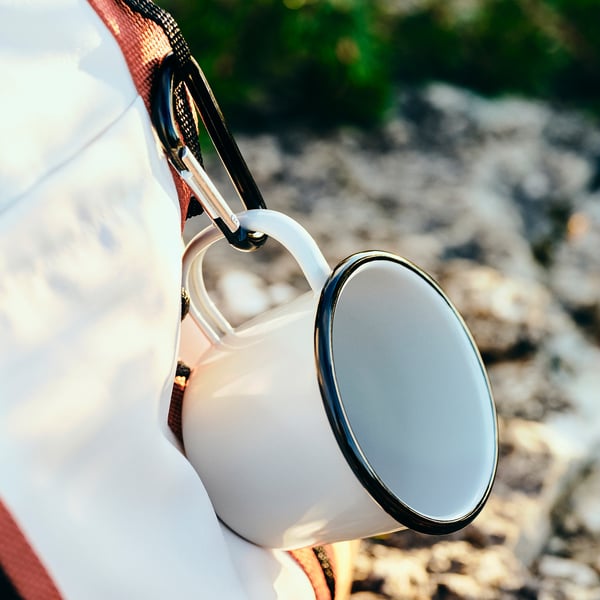 A white enamelled steel mug securely attached to a carabiner on a backpack, shown outdoors.