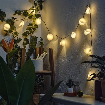 String of solar-powered flower lights illuminating a wall, surrounded by lush green plants in pots.