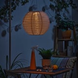 Cosy outdoor setting with a SOLVINDEN solar-powered lantern casting warm light over a table with plants, drinks, and an orange.
