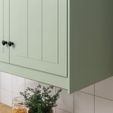 Green kitchen cabinets with dark knobs and plant.