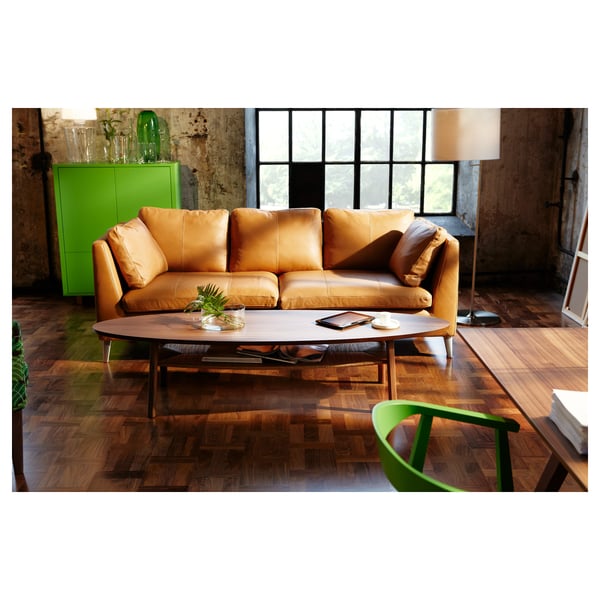 A modern living room with a brown leather sofa, a dark oval wooden coffee table with a shelf, and green decor items including a cabinet and chair.