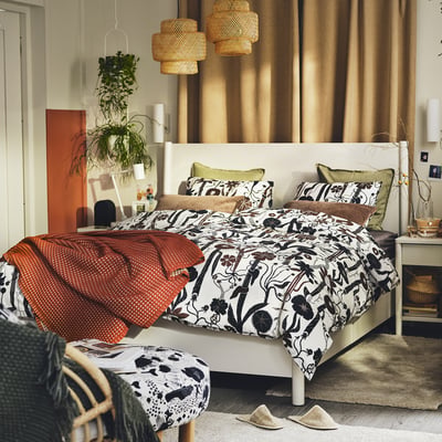 Bedroom with white rug, black floral bedding, green pillows, red blanket, plants, wicker lamps, slippers.