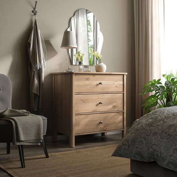 Bedroom with light wood STOREMOLLA dresser, mirror, lamp, and green plant. Gray chair and curtains.
