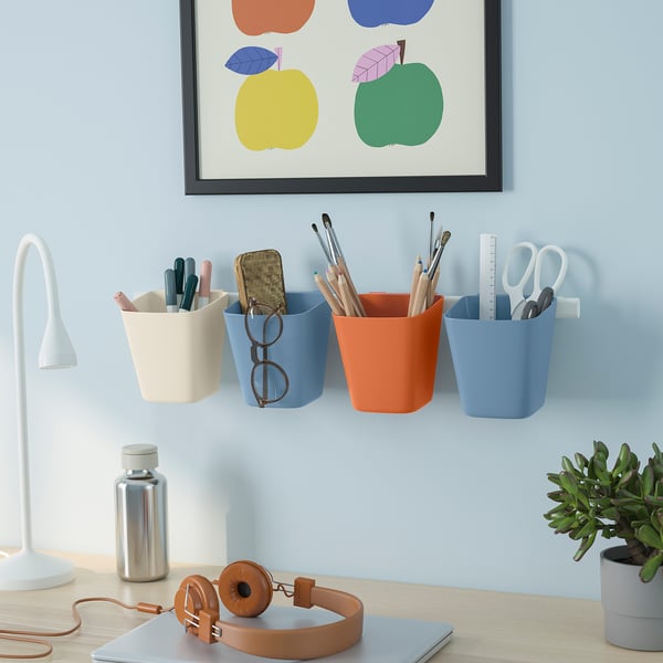 Three colorful SUNNERSTA organizers on blue wall, each with office supplies. Above desk with lamp, bottle, headphones, plant, and apple frame.