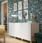 Modern living room SUTTERVIKEN white cabinet with gold accents against dark floral wallpaper, adorned with framed art and a table lamp.