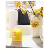 A yellow SVARTOXBÄR candle in a frosted glass with circular designs sits on a wooden table next to a vase with yellow flowers.