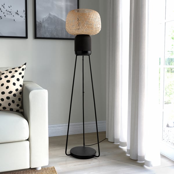 SYMFONISK wi-fi speaker lamp in black - modern, tall, bamboo shade, stands beside white couch, lit window nearby.