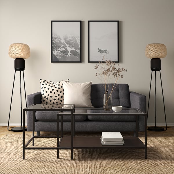 Modern living room with SYMFONISK wi-fi speaker floor lamp on both sides of grey couch. Framed art on wall, glass coffee table with books.