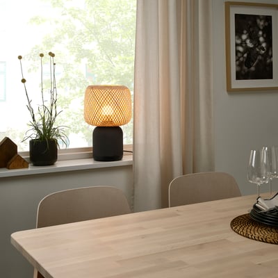 Small bamboo shade on speaker lamp sits near window, between curtains and chairs, beside a potted plant.