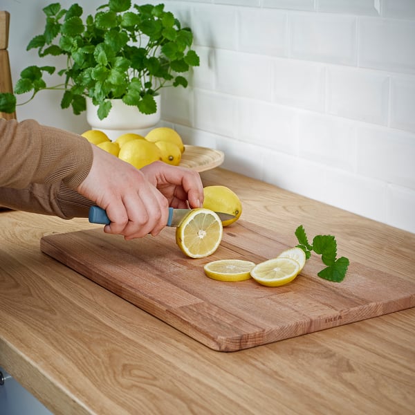 Person slices lemon on wooden cutting board with knife. Lemons and herbs nearby, on wooden countertop.