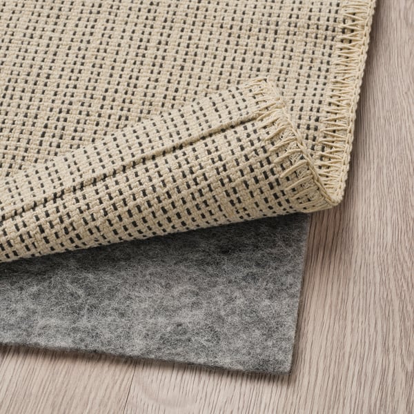 A beige woven rug with dark accents, placed on a gray non-slip pad on a wooden floor.