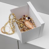 A white TJENA storage box filled with jewelry, placed on a gray surface. The box is open, showcasing its function for storing small items neatly.
