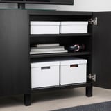Black cabinet with open doors reveals organized storage: two white TJENA boxes, stacked notebooks, and headphones.