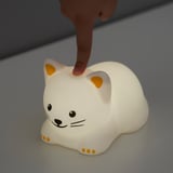 A hand touches a TÖVÄDER night light shaped like a white cat with orange spots and ears, emitting a soft glow.