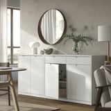 Modern dining room with white TONSTAD cabinet, round mirror, and wooden chairs. Greenery in vase, lamps, and soft lighting.