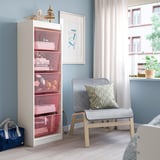 This image shows a white TROFAST bookshelf with pink mesh storage boxes, surpassing a grey modern chair by a window.