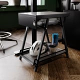 Black TROTTEN storage table with lockable drawer and mesh shelf holds various items, positioned on wooden floor in office.