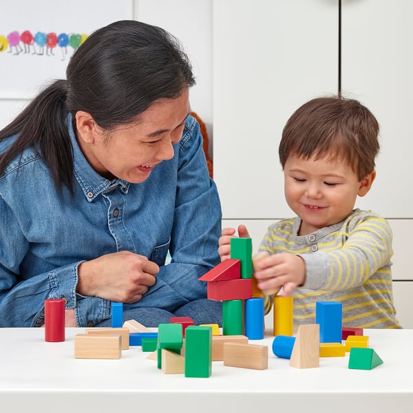 A young child and an adult play with colorful wooden blocks on a white table. The child is stacking them, learning shapes and colors.
