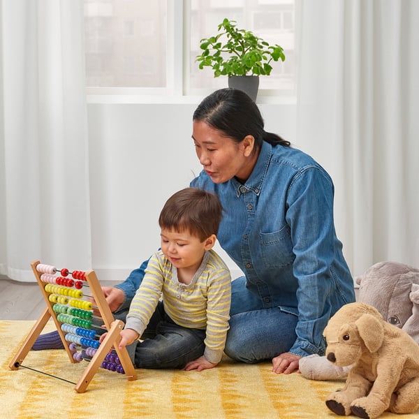A woman and child play with a colorful, foldable abacus on a yellow rug, surrounded by soft toys.