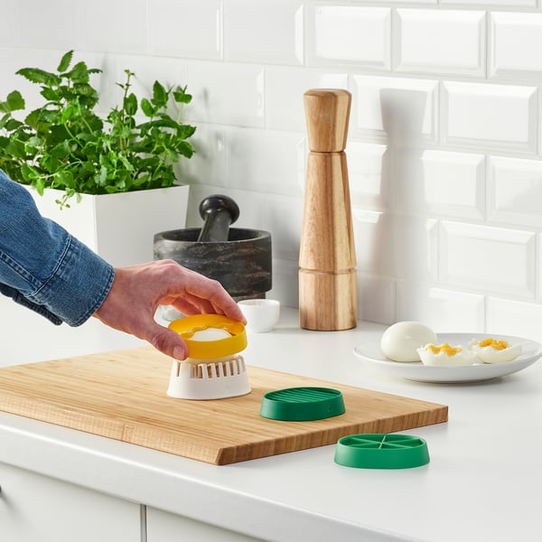 Person uses colorful egg slicer on wooden board. Modern kitchen with plant.