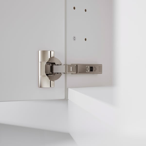 A modern kitchen hinge, adjustable in three directions, mounted on a white cabinet. Ideal for precise door alignment and easy removal for cleaning.