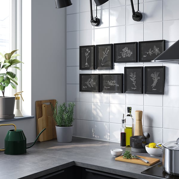 Modern kitchen with white walls, dark counters, VÄXBO art, green watering can, and plant pots.
