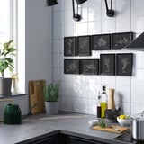 Modern kitchen with white walls, dark counters, VÄXBO art, green watering can, and plant pots.