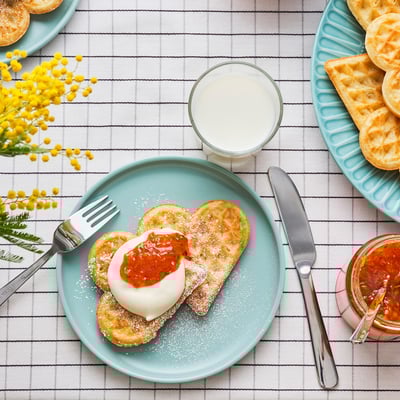 Heart-shaped waffles with sugar, cream, jam on turquoise plate with utensils and flowers.